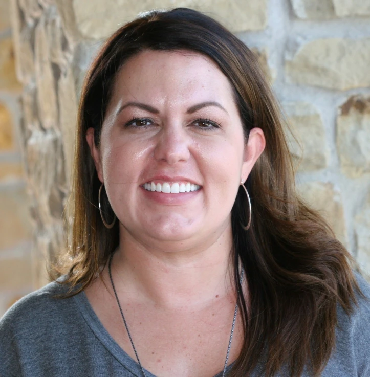 Smiling woman with long brown hair wearing hoop earrings and a gray top standing in front of a stone wall.