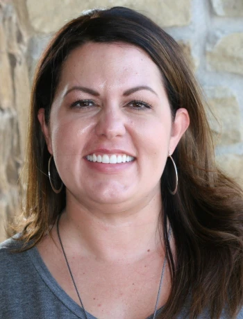 Smiling woman with long dark hair, hoop earrings, and a gray top standing in front of a stone wall.