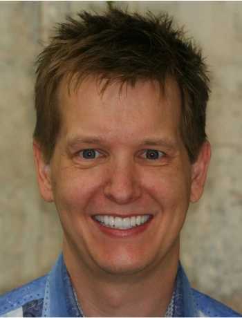 Smiling man with short spiky brown hair wearing a blue patterned shirt against a blurred background.