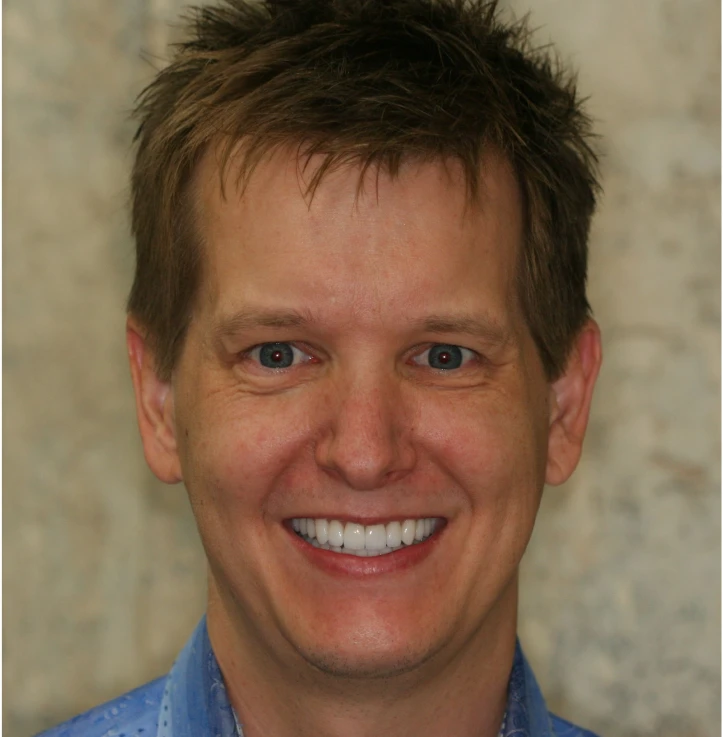 Smiling man with short brown hair wearing a blue shirt in front of a blurred background.