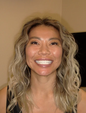 Smiling woman with long wavy blonde hair and tan skin, wearing a black top against a beige background.