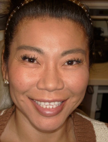 Close-up of a smiling woman with long eyelashes and pearl earrings wearing a brown and white sweater.