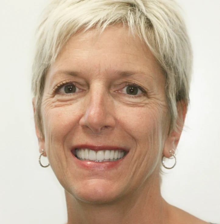 Smiling middle-aged woman with short blonde hair and hoop earrings against a plain light background.