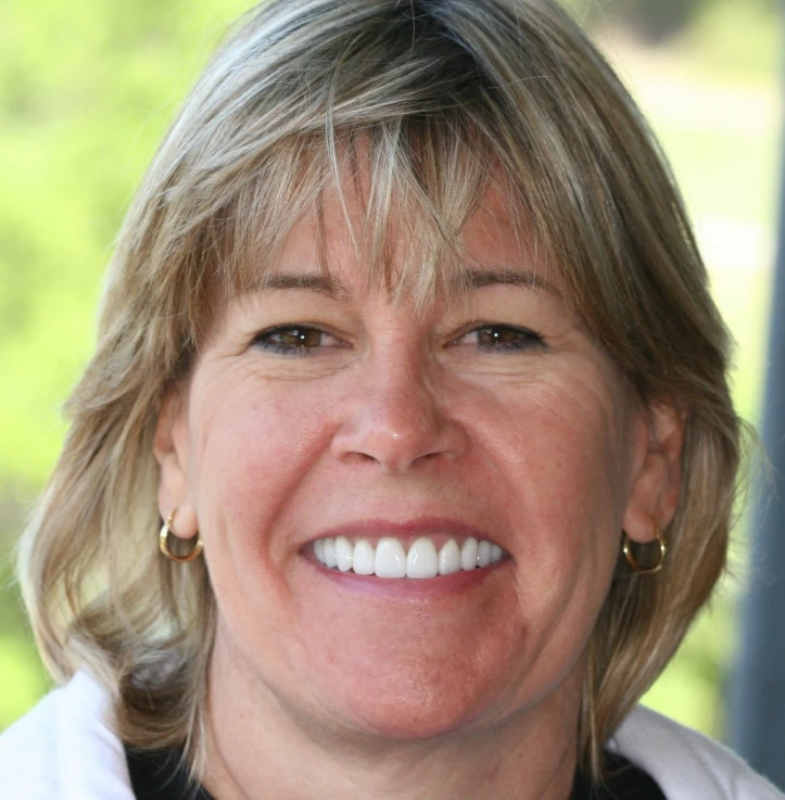 Smiling woman with short blonde hair and hoop earrings in a bright outdoor setting.