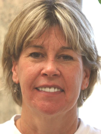 Close-up of a smiling middle-aged woman with short blonde hair, wearing earrings and a white top.