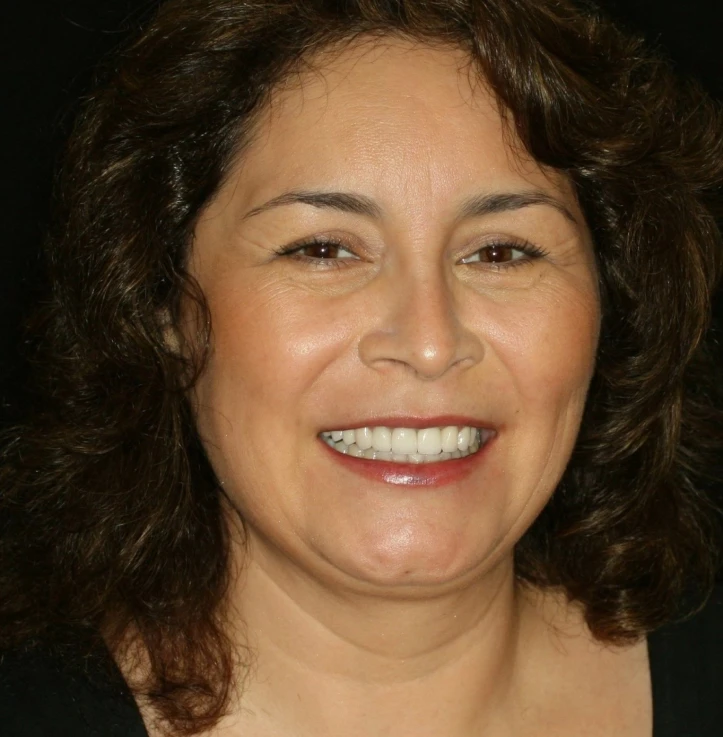 Smiling middle-aged woman with curly dark hair and light makeup against a black background.