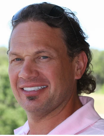 Smiling man with curly dark hair wearing sunglasses on his head and a light pink polo shirt outdoors.