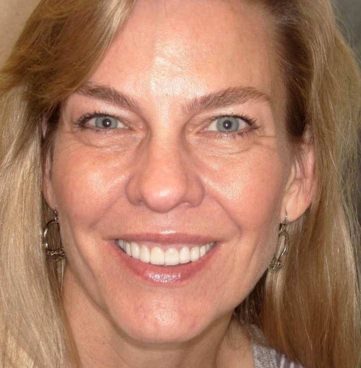 Close-up of a smiling middle-aged woman with blonde hair and blue eyes wearing silver earrings.