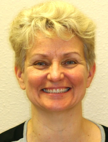 Smiling woman with short curly blonde hair and light skin wearing a black and gray top against a plain beige background.