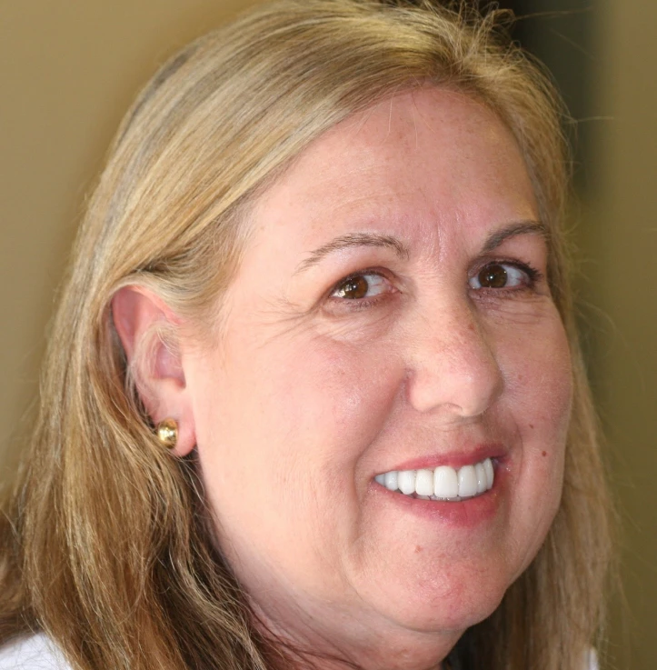 Close-up of a smiling middle-aged woman with blonde hair and gold earrings against a neutral background.
