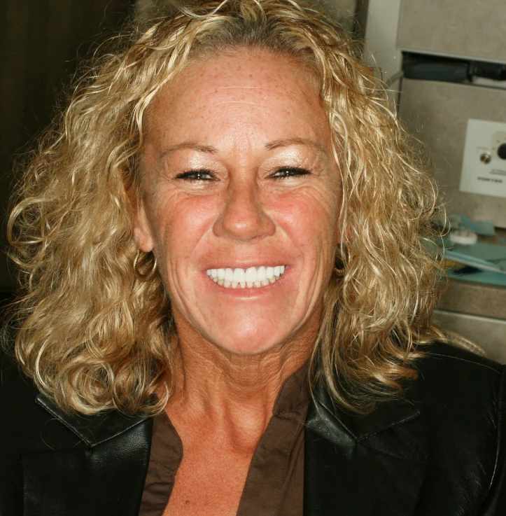 Smiling middle-aged woman with curly blonde hair wearing a black jacket and brown shirt.