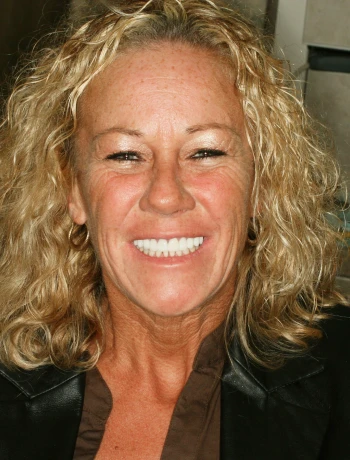 Smiling woman with curly blond hair wearing a black jacket and brown top.