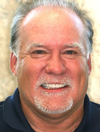 Smiling middle-aged man with gray hair and a short gray beard wearing a dark collared shirt.