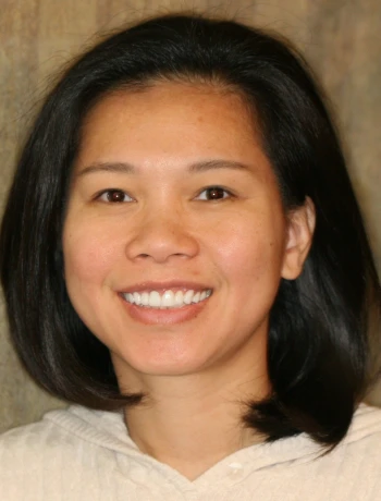Smiling woman with straight black hair wearing a light-colored sweater, in front of a neutral background.