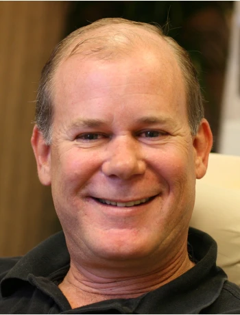 Smiling middle-aged man with short hair wearing a black shirt seated indoors.