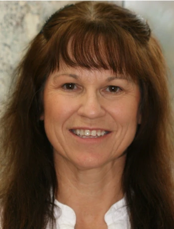Smiling middle-aged woman with long brown hair and bangs wearing a white top.