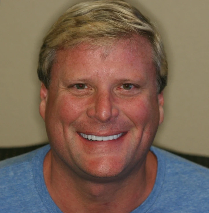 Smiling man with short blonde hair wearing a blue shirt against a plain background.