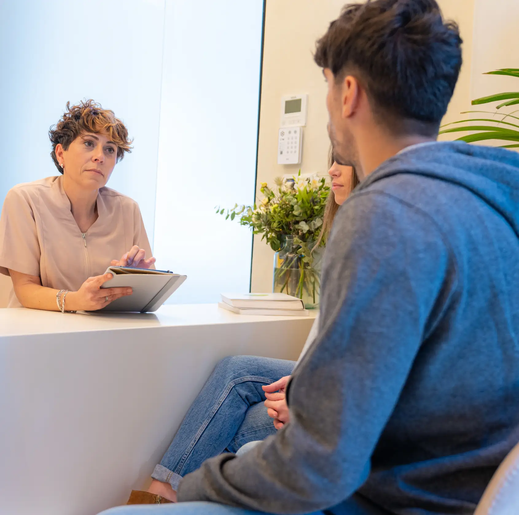 Woman with short curly hair holding a tablet, attentively talking to a couple seated across from her in a bright office.