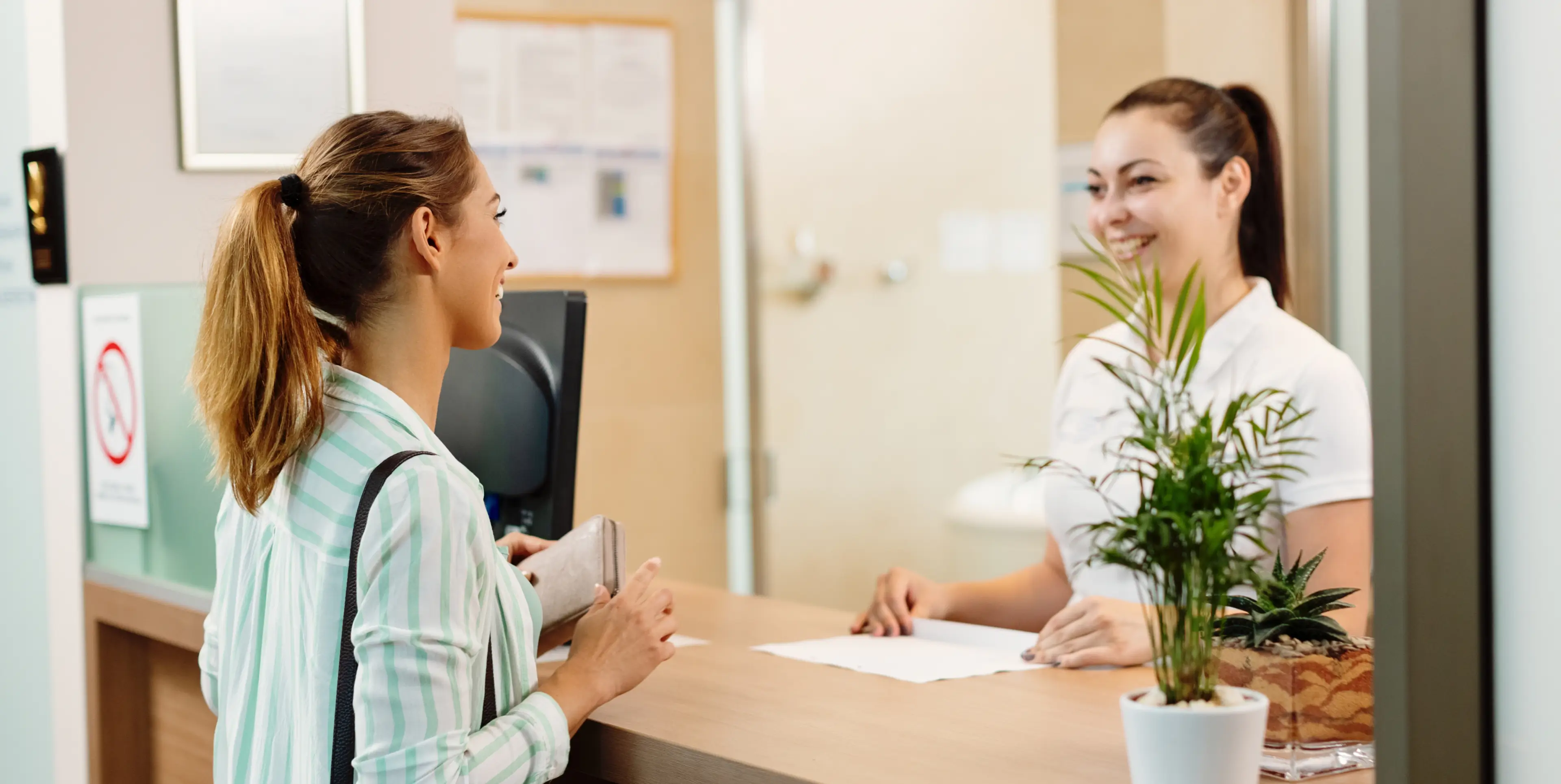 A woman with a ponytail and striped shirt talking to a smiling receptionist at a desk with potted plants.