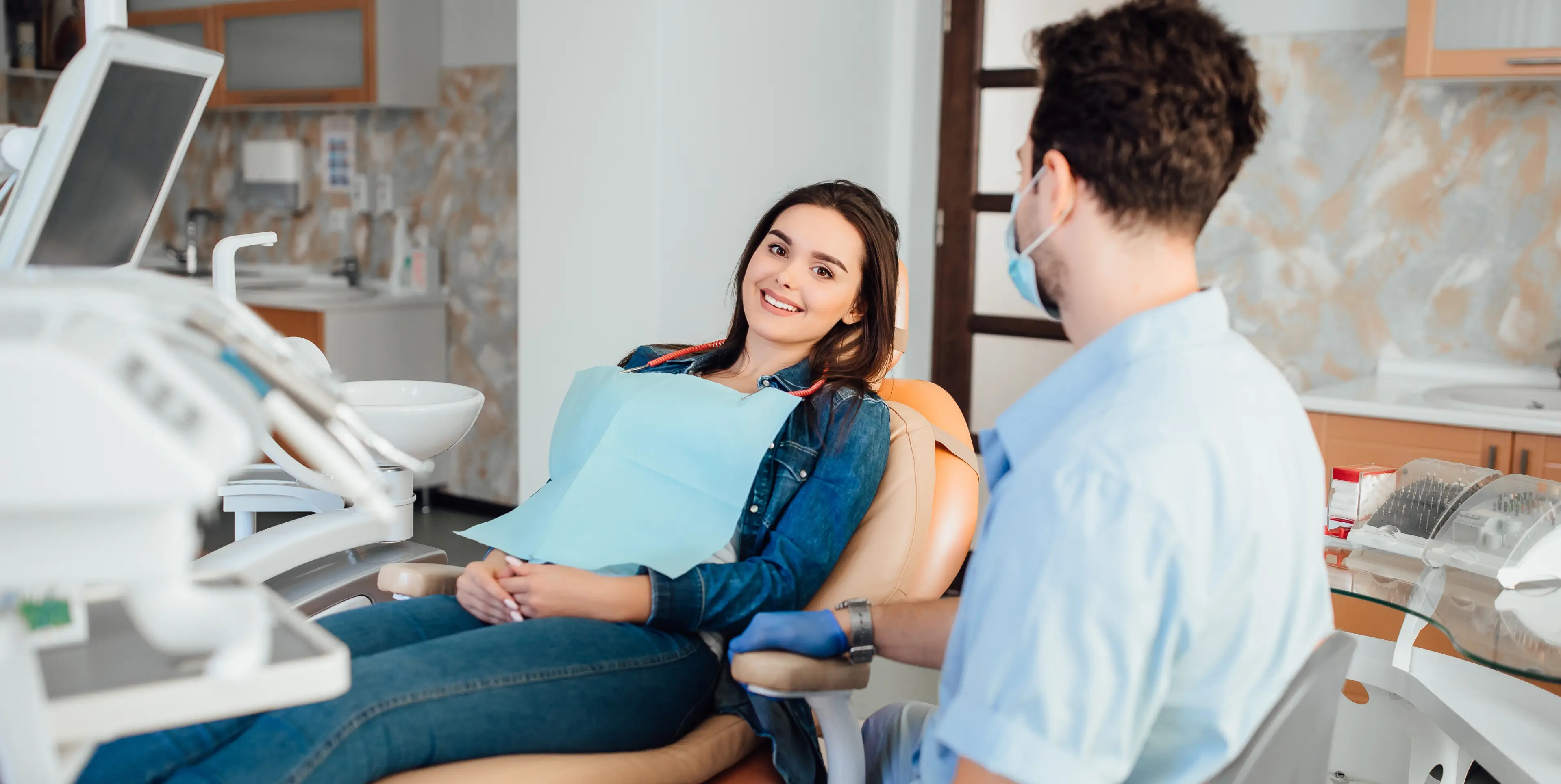 Smiling woman in a dental chair wearing a blue bib, talking to a masked dentist in a light blue uniform.