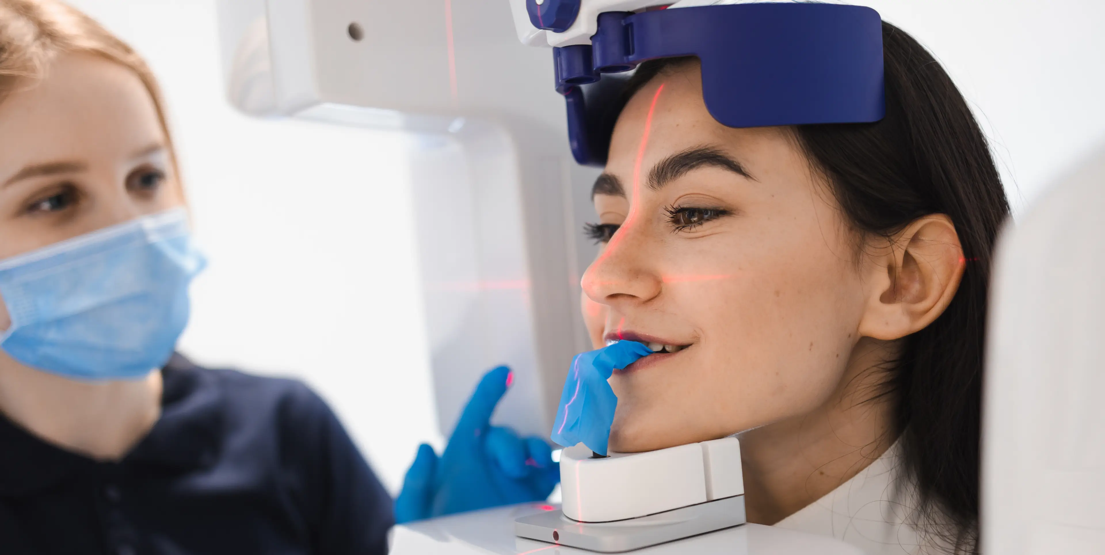 Woman undergoing a 3D dental scan with a blue bite piece and red laser guide, supervised by a masked dental professional wearing blue gloves.