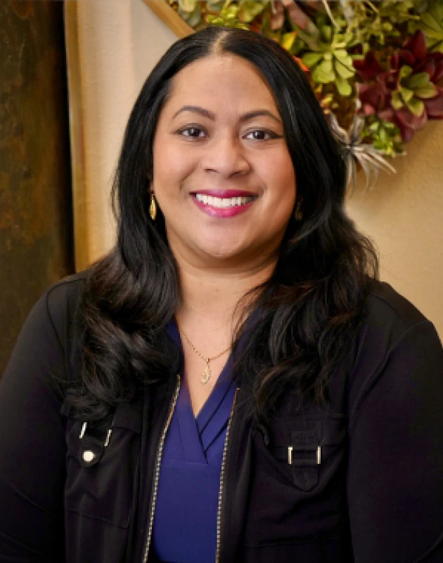 Smiling woman with long black hair wearing a dark jacket and blue top, standing indoors with green plants in the background.