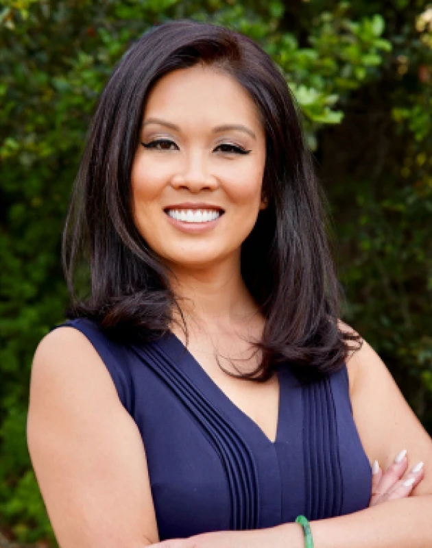 Smiling woman with shoulder-length dark hair wearing a sleeveless navy blue top standing outdoors with arms crossed.