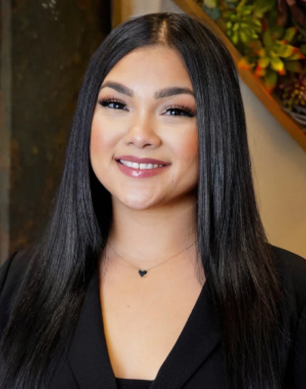 Portrait of a smiling woman with long straight black hair wearing a black top and a small heart-shaped necklace.