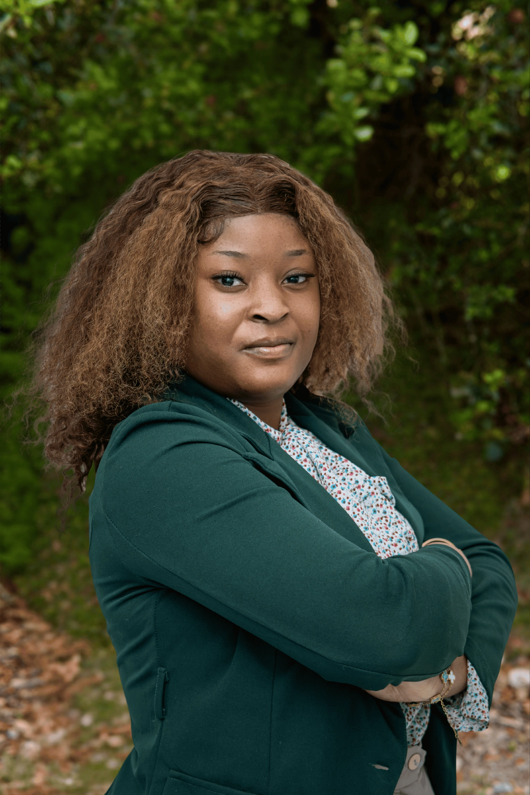Confident woman with curly brown hair wearing a dark green blazer and floral blouse, standing outdoors with arms crossed.