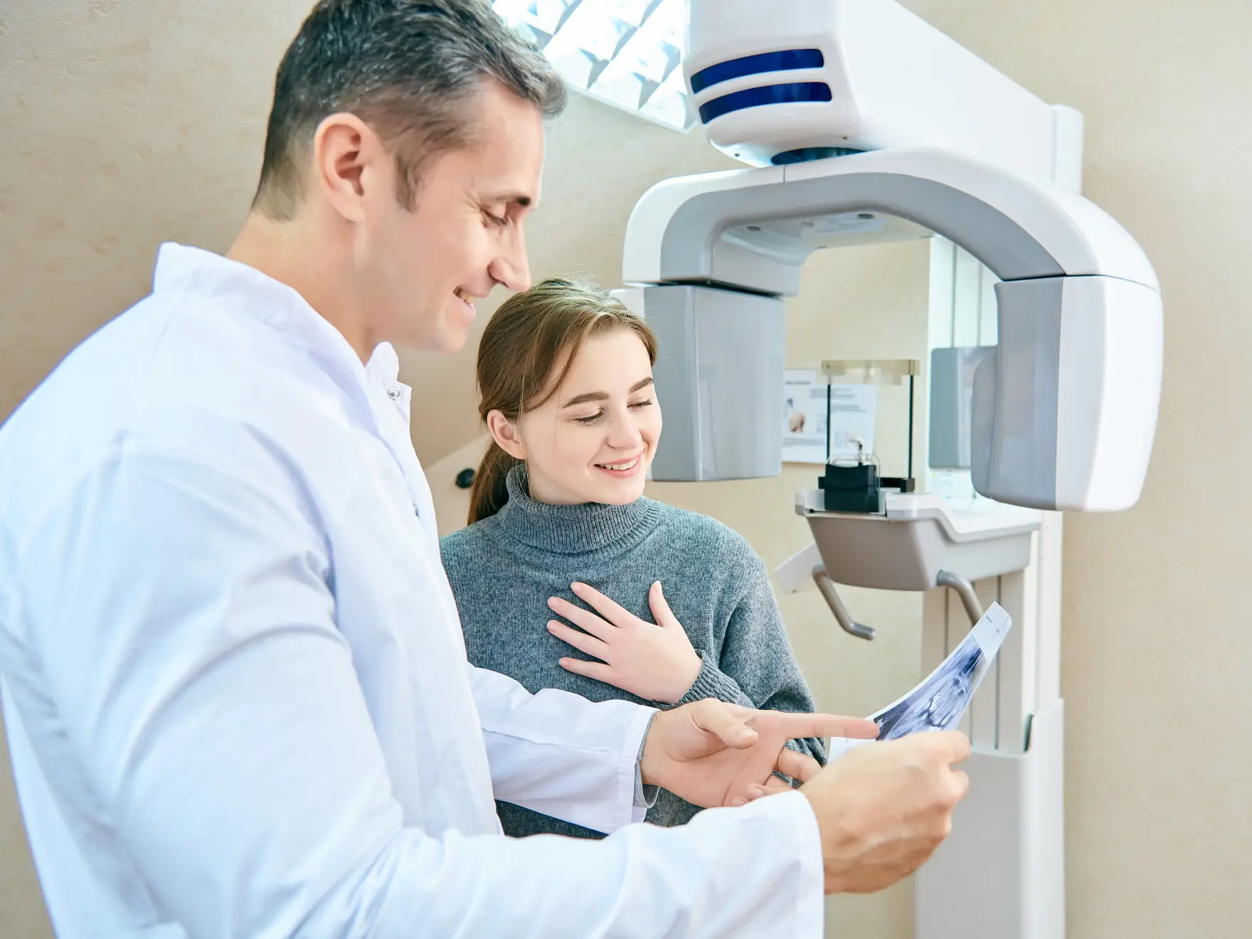 Dentist in a white coat showing a dental X-ray to a smiling female patient next to a panoramic dental X-ray machine.