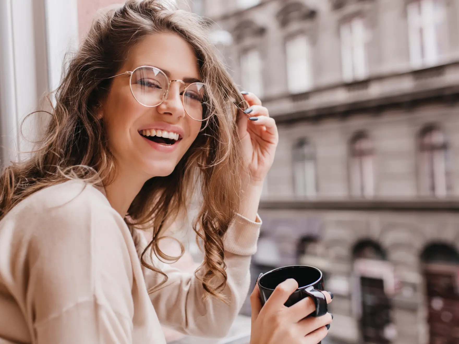 Smiling woman with curly hair and glasses holding a black mug by a window with blurred city buildings in the background.