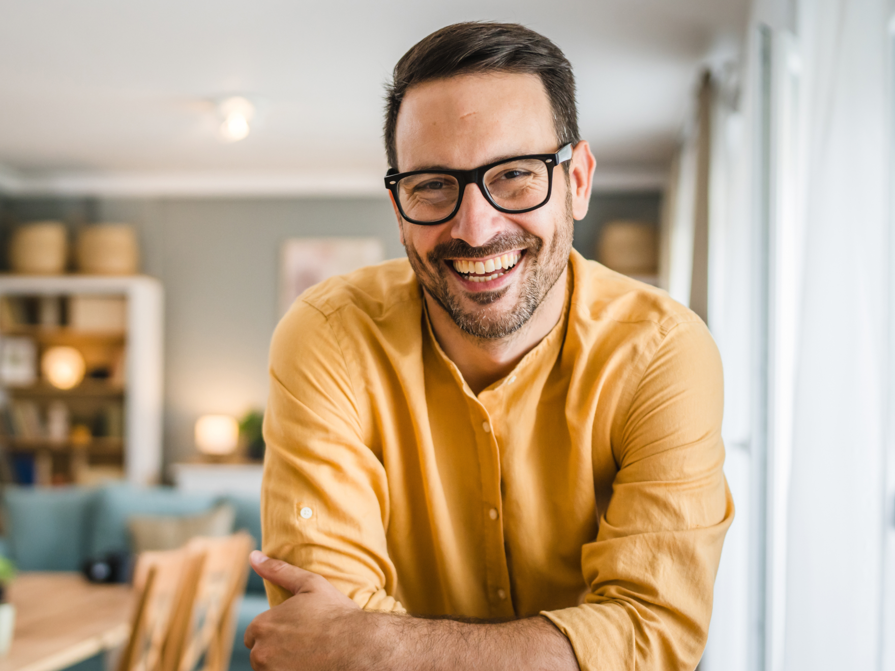Smiling man in glasses and mustard-yellow shirt leaning on wooden table in a bright living room.