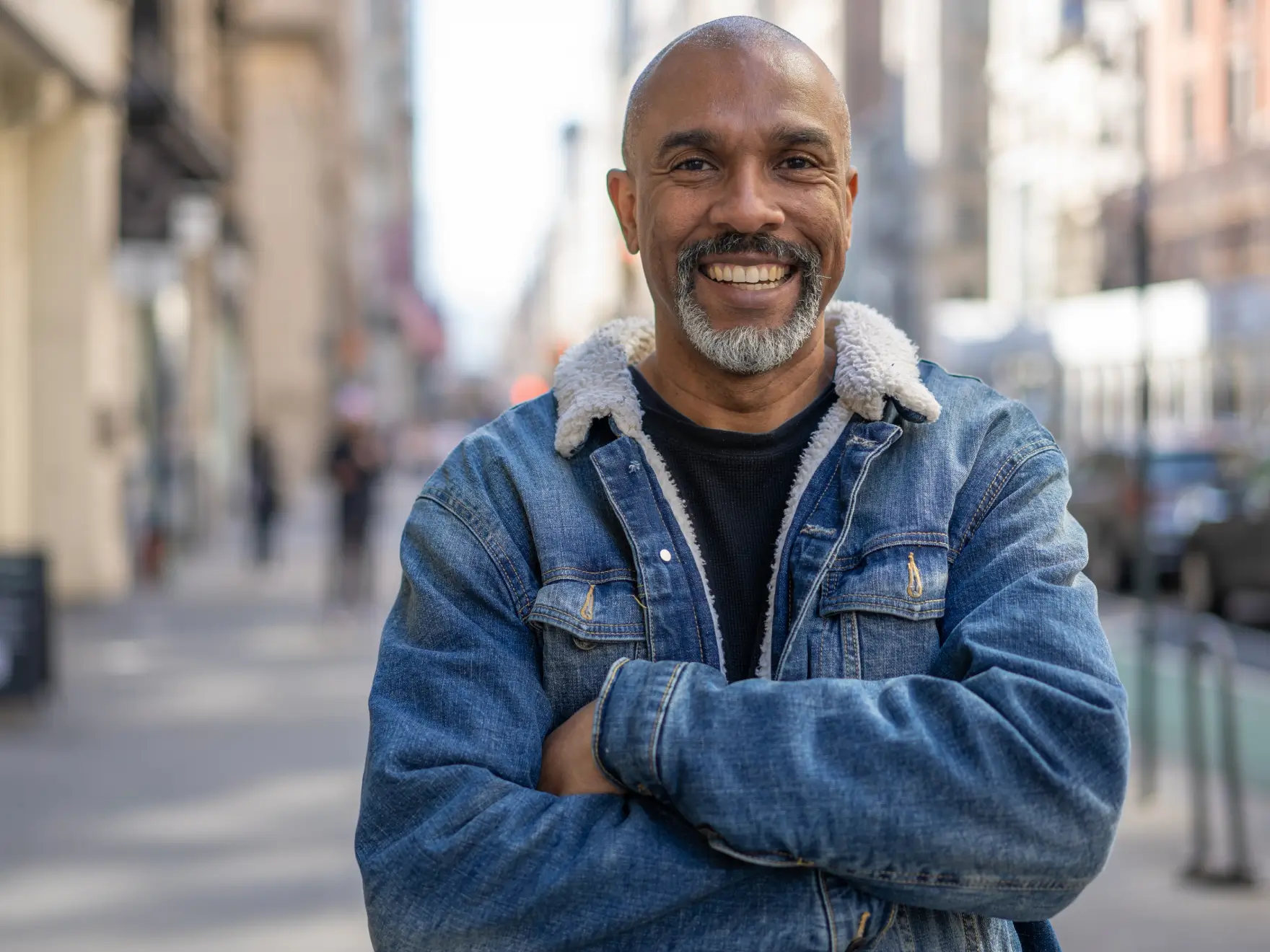 Smiling middle-aged man with gray beard wearing a blue denim jacket standing outdoors on a city street.