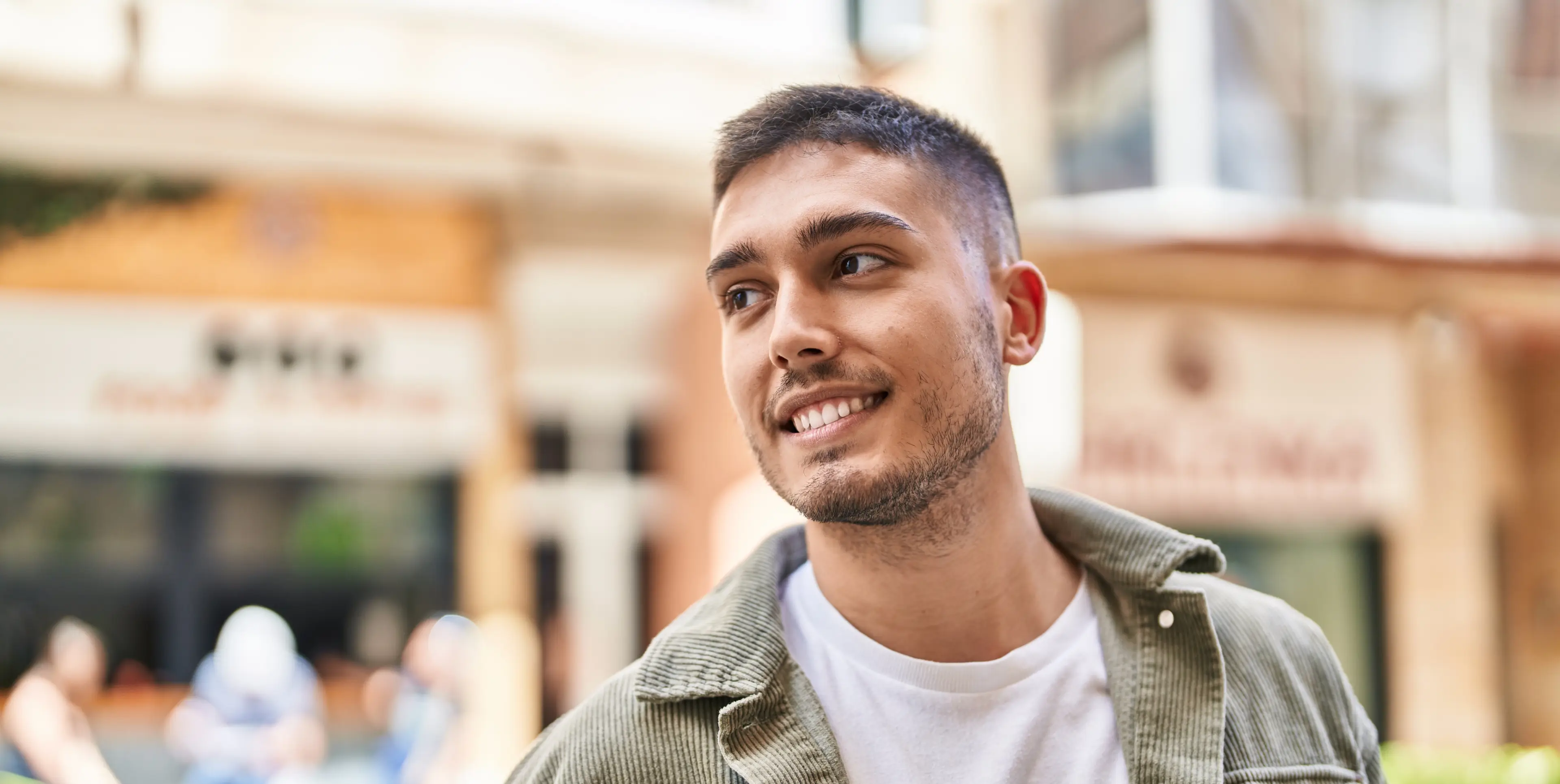 Young man with short dark hair and beard smiling and looking to the side outdoors with blurred background.