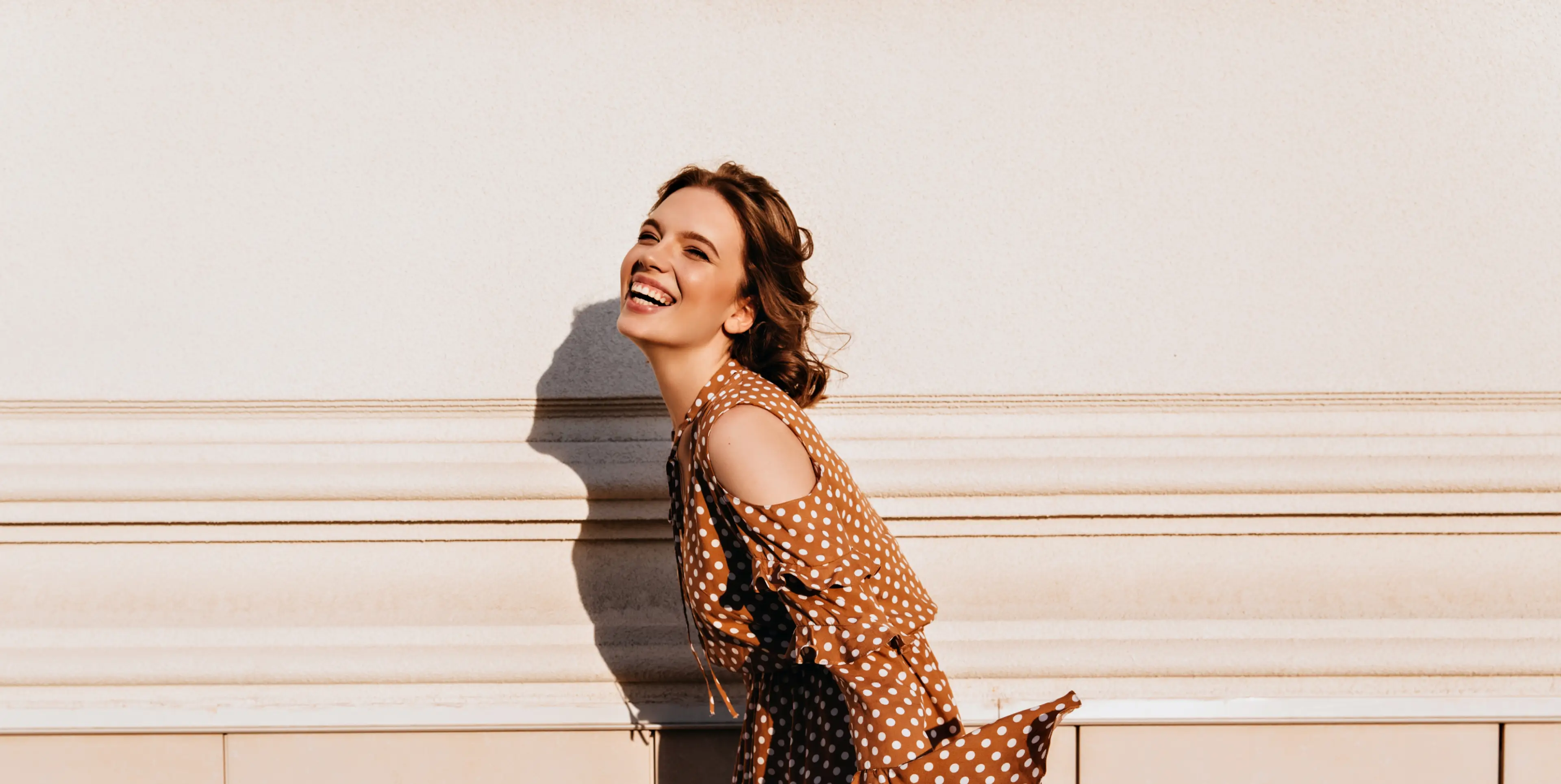 Smiling woman in a brown polka dot dress leaning forward against a beige wall with shadows.