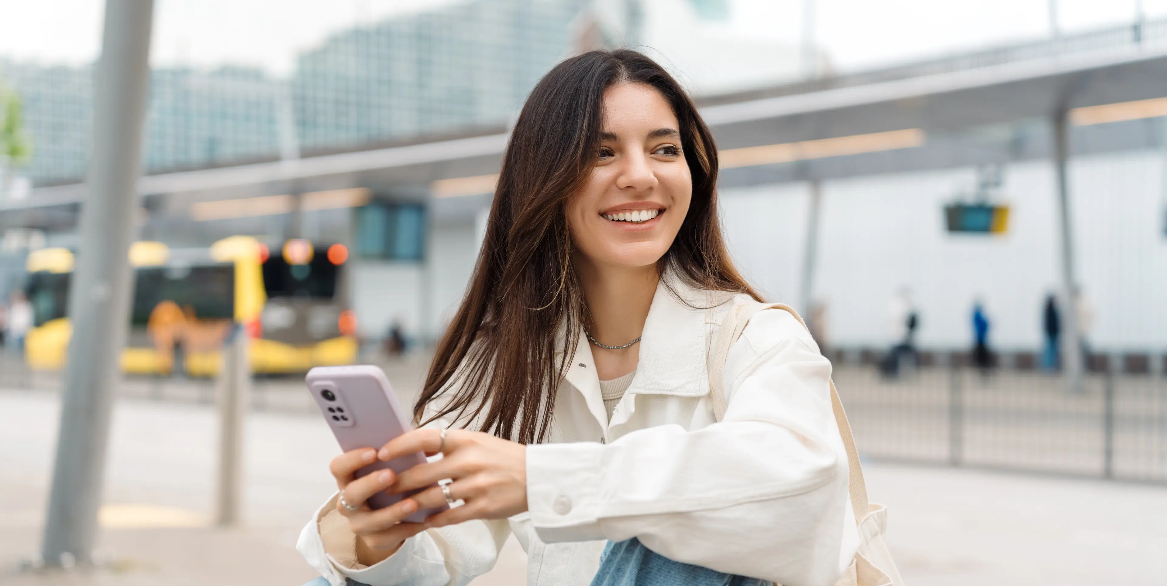Smiling young woman with long dark hair in a white jacket holding a smartphone outdoors near a bus stop.