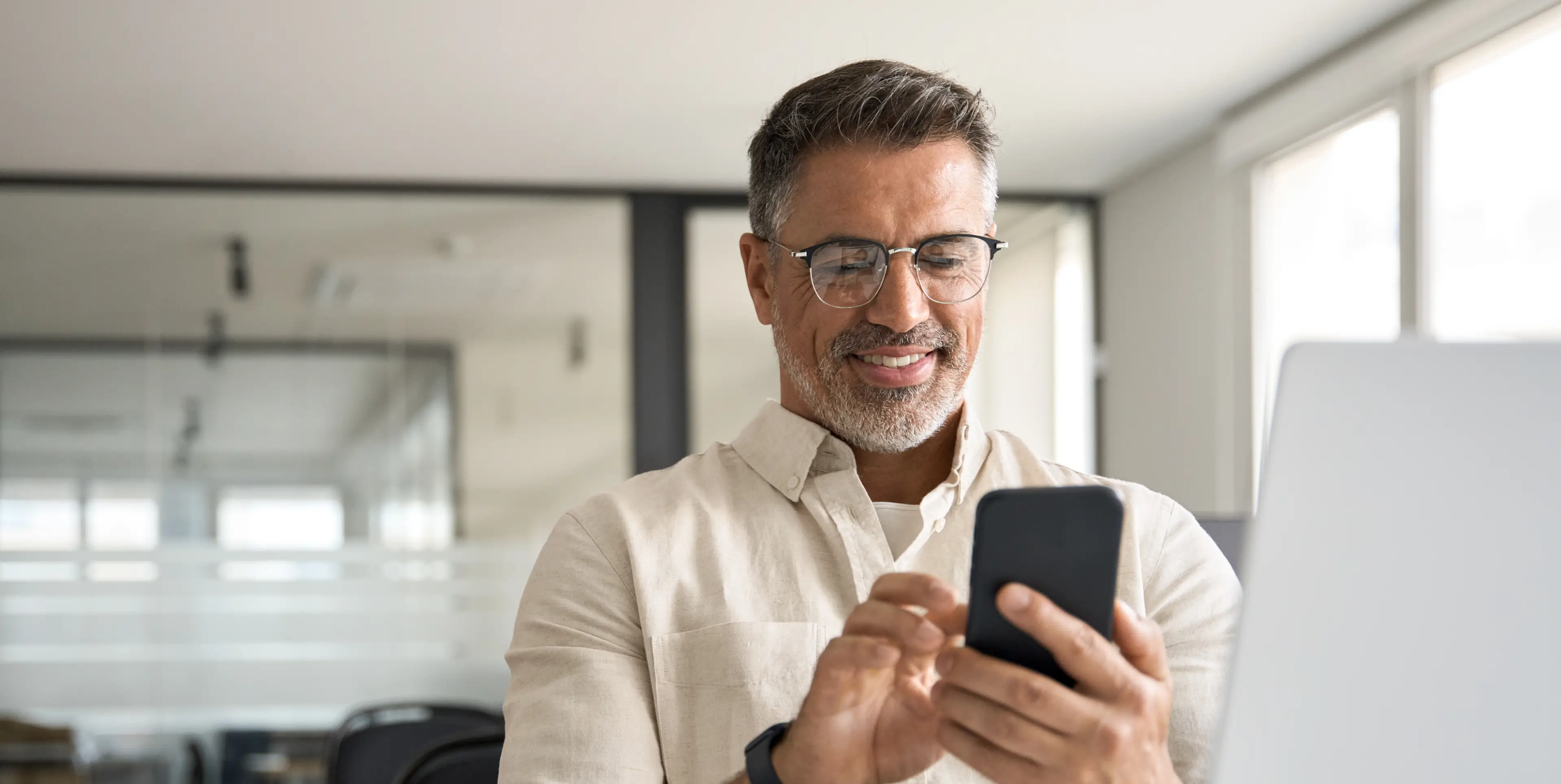 Smiling middle-aged man with glasses using smartphone in a bright office.
