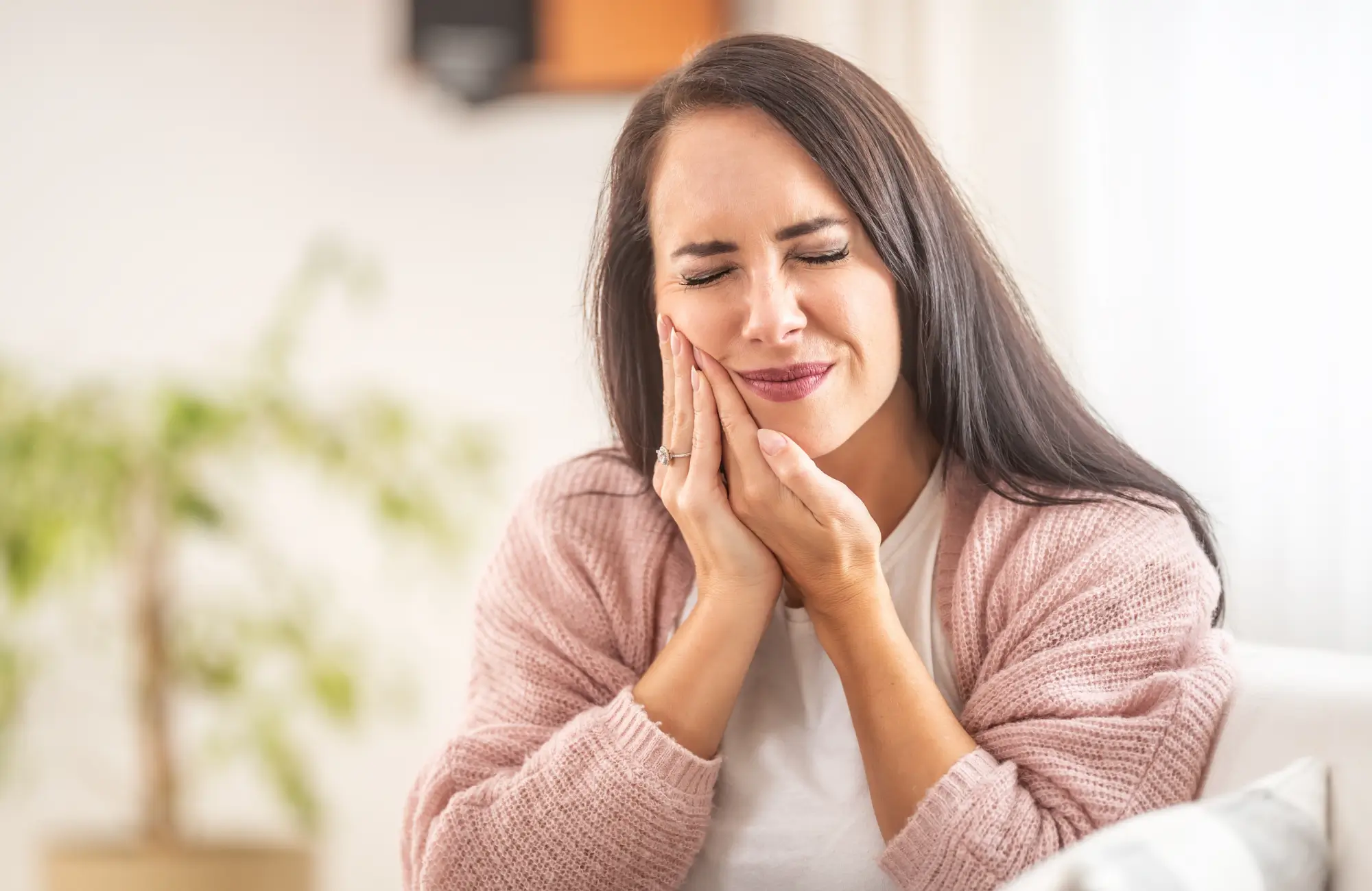Woman with long dark hair wearing a pink cardigan, holding her jaw with a pained expression, indicating toothache or jaw pain.