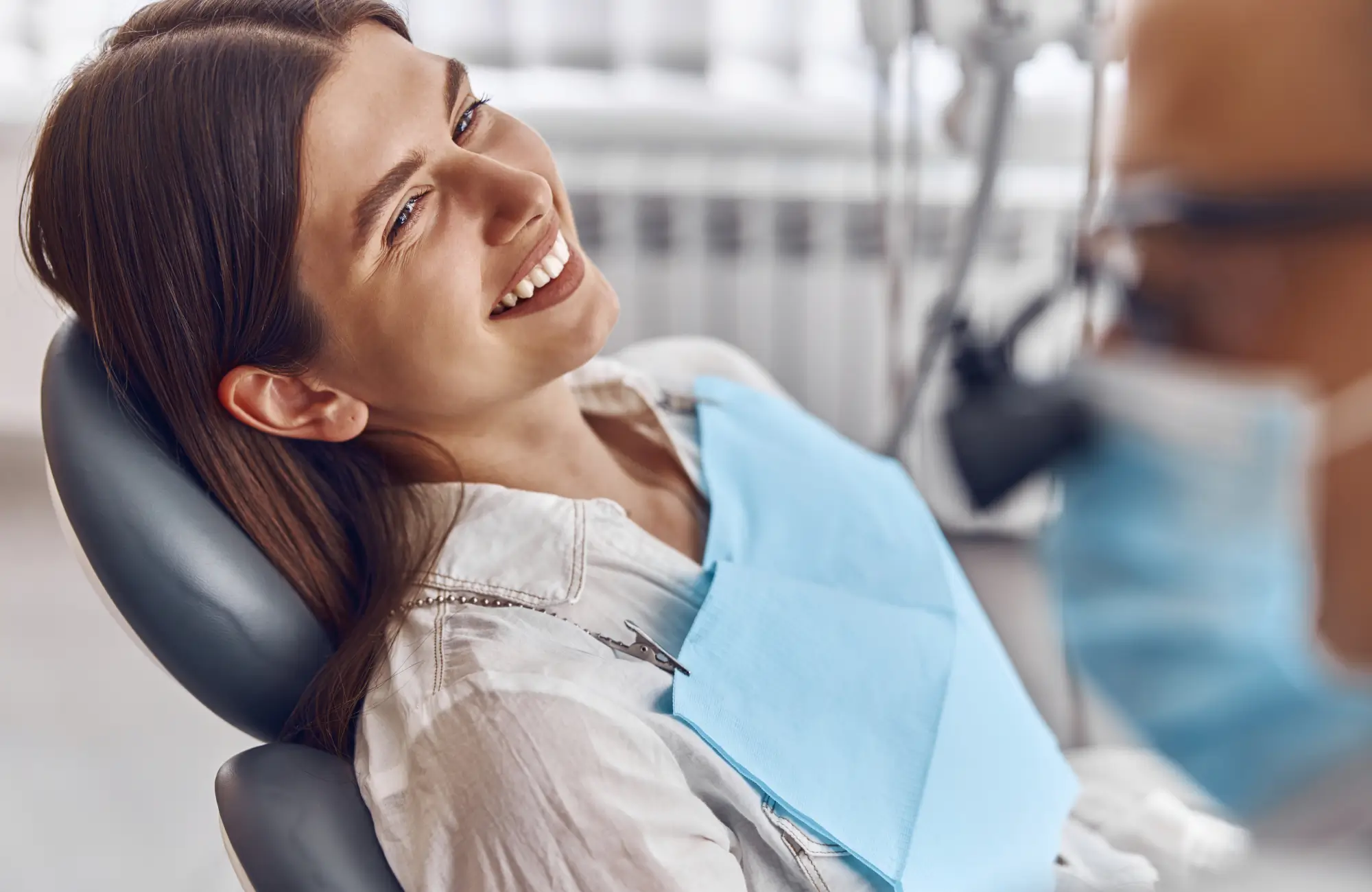 Smiling woman reclined in a dental chair with a blue bib, facing a dentist wearing a mask.