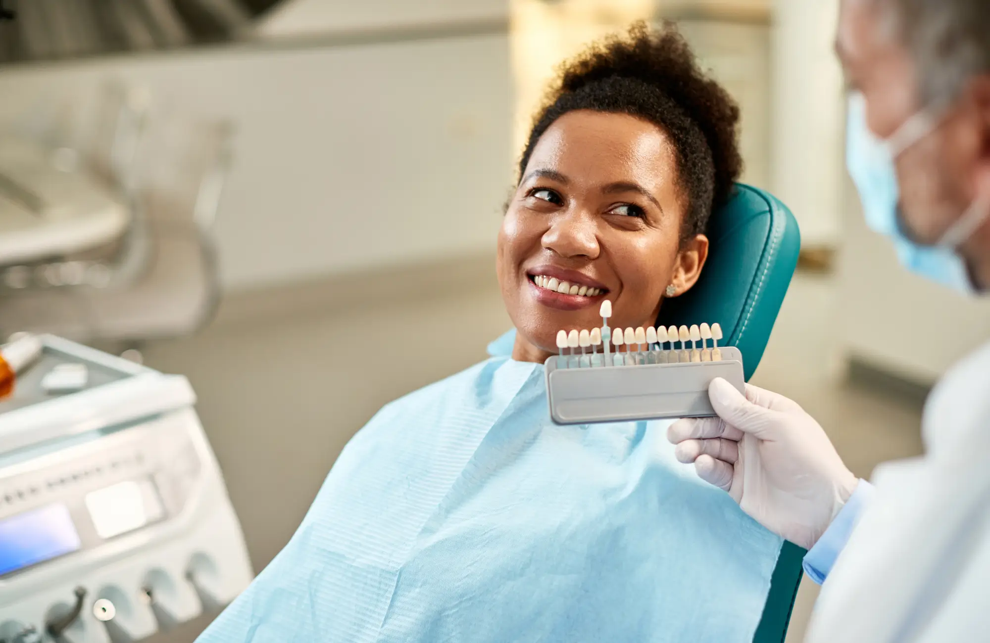 A female patient smiling in a dental chair while a dentist holds a shade guide of dental veneers near her teeth.