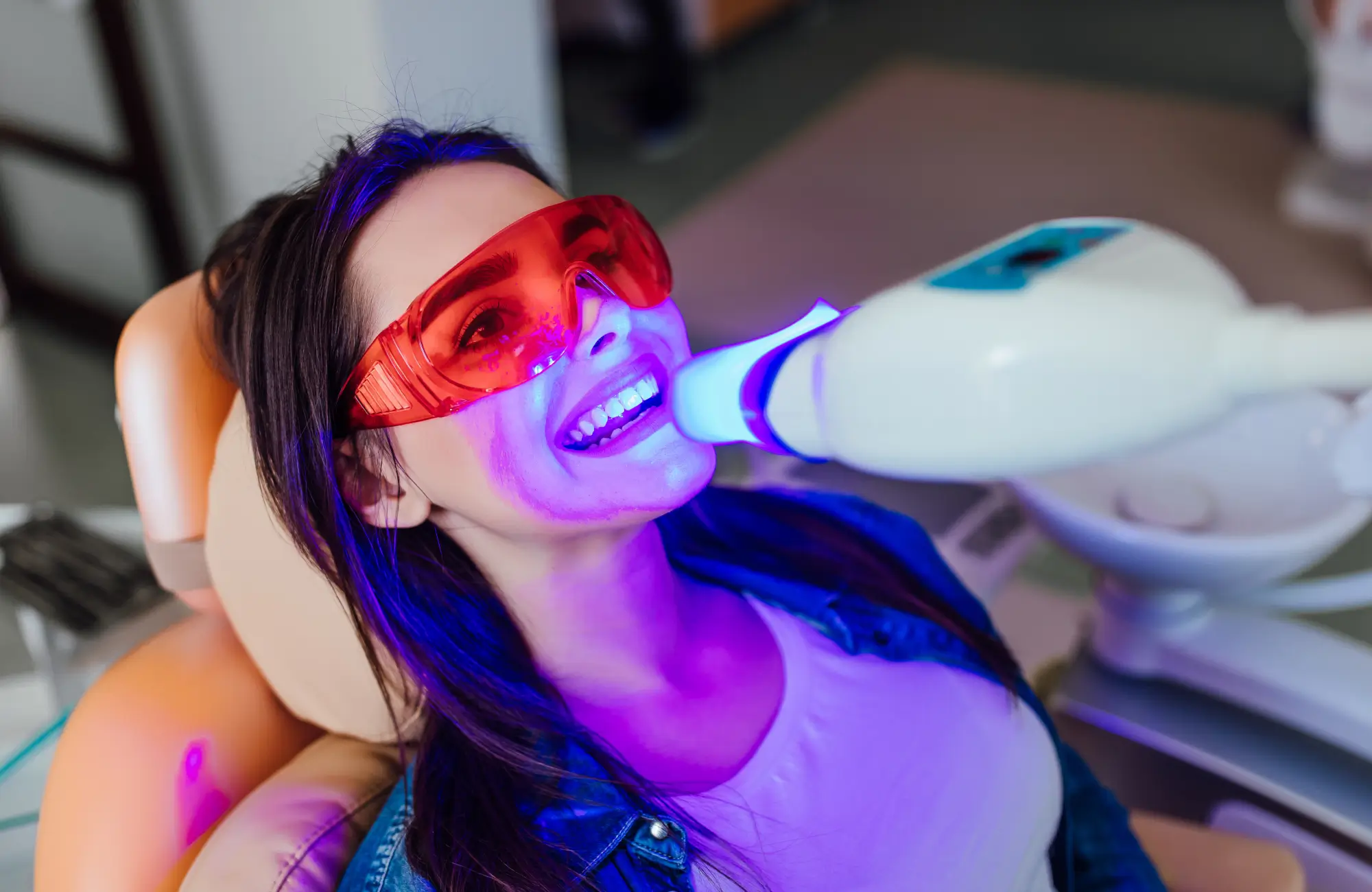 Woman wearing red protective glasses undergoing blue light teeth whitening treatment in dental chair.