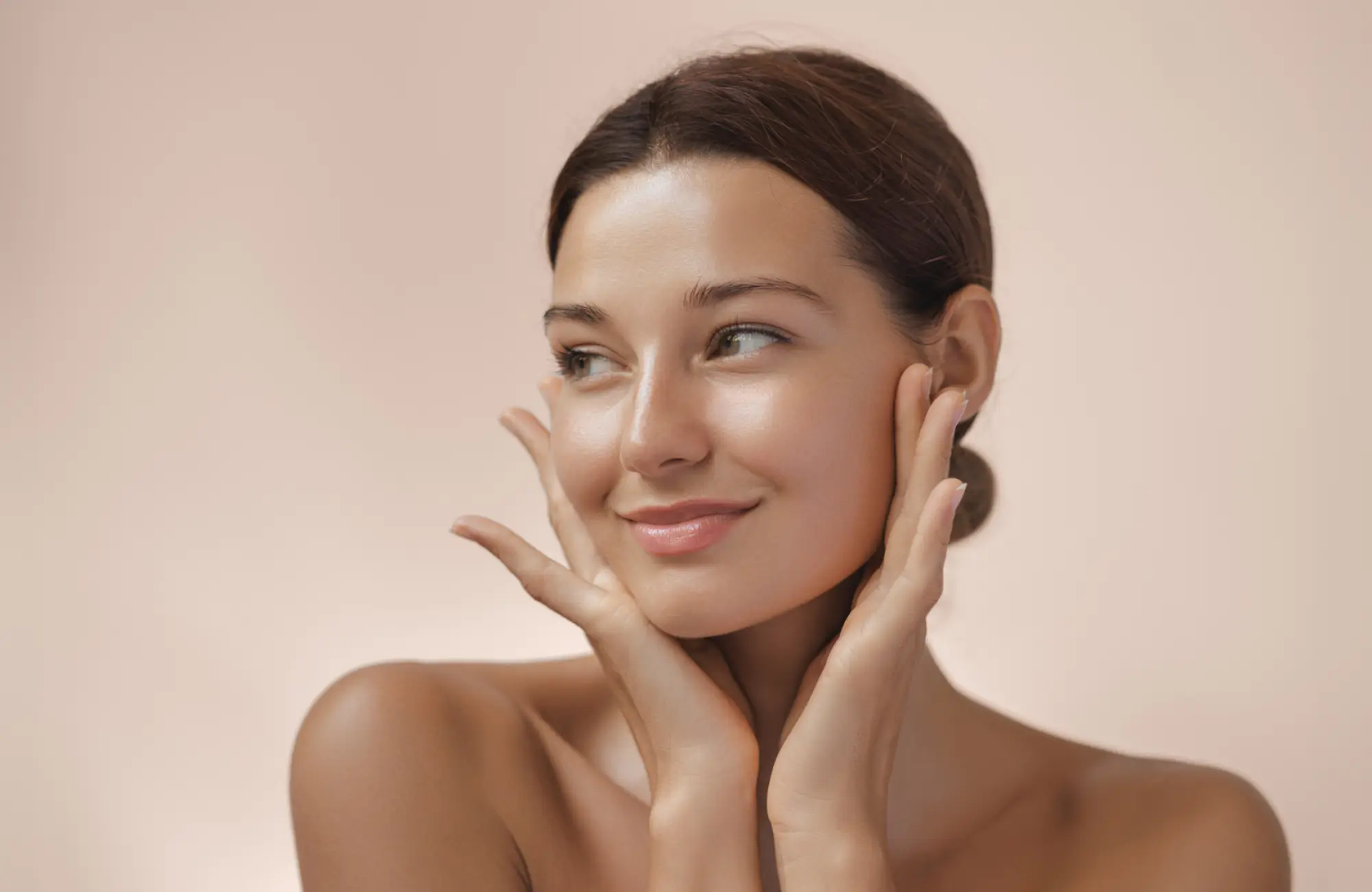 Smiling woman with clear skin gently touching her face against a soft beige background.