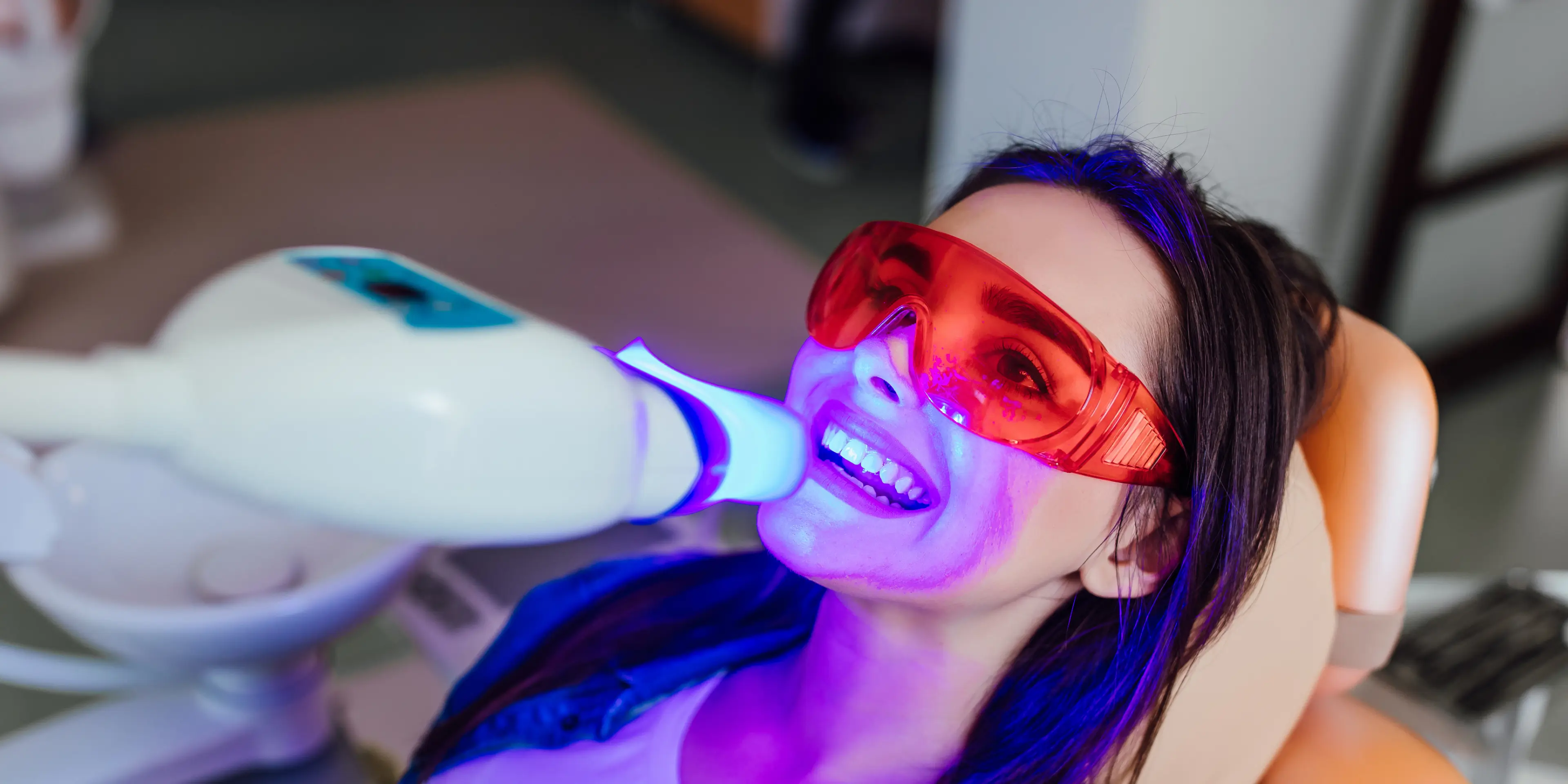 Woman wearing red protective glasses receiving blue light teeth whitening treatment at dental clinic.