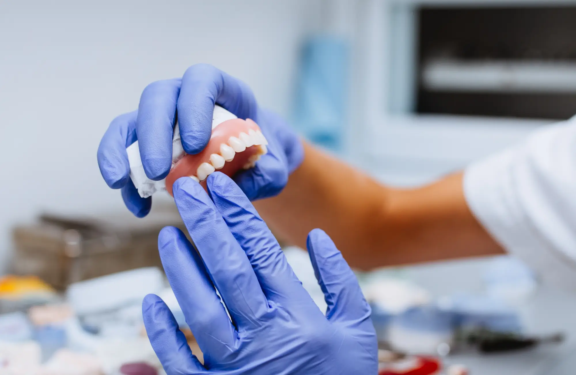 Hands in blue gloves holding and inspecting a dental prosthesis with artificial teeth.