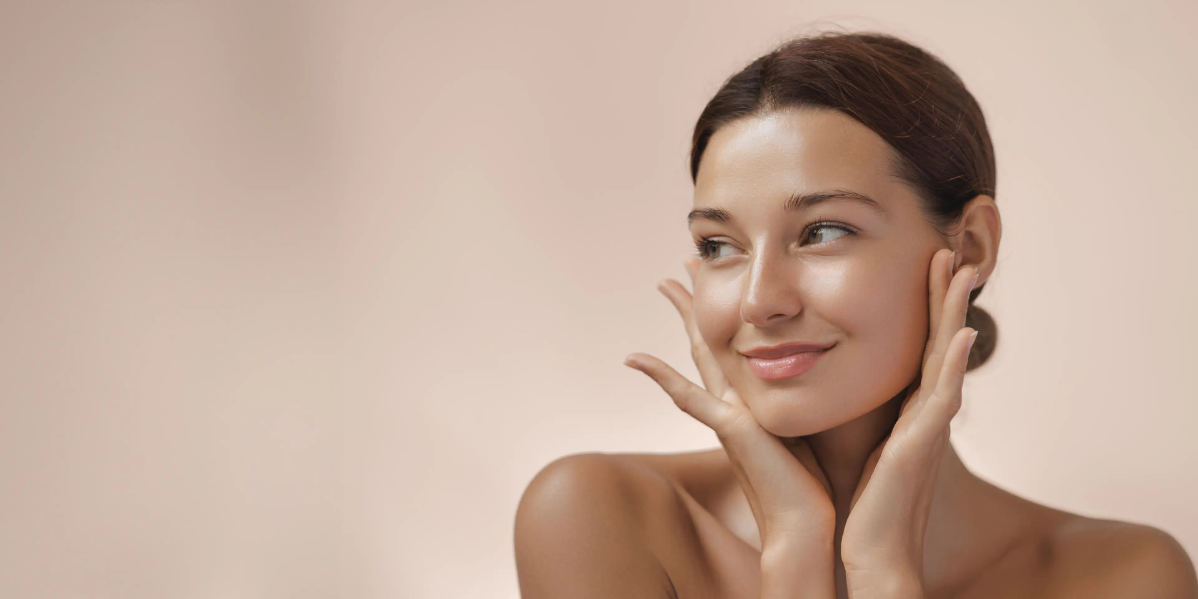 Smiling young woman with clear skin gently touching her face against a soft beige background.