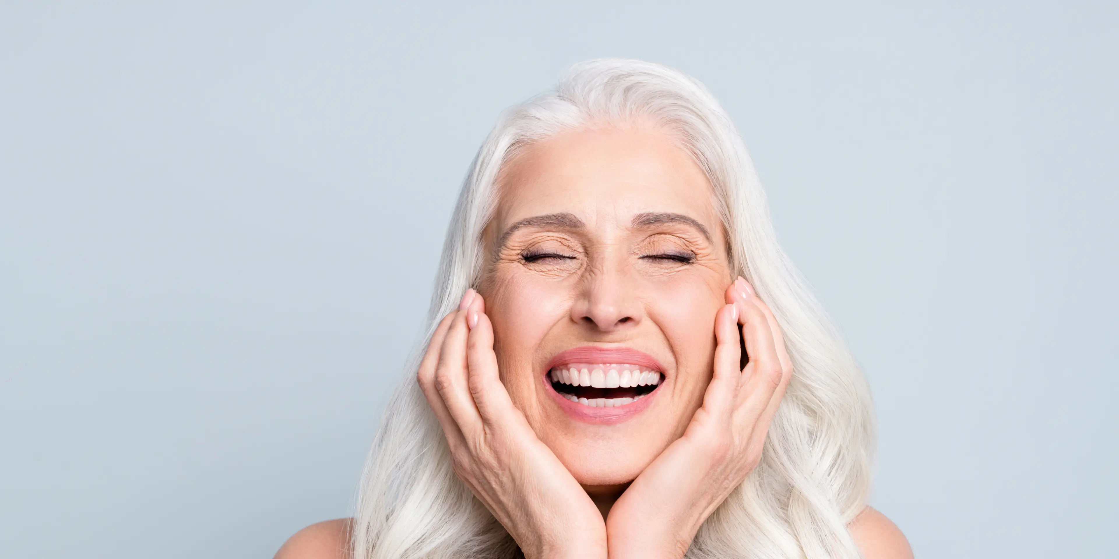 Close-up of a joyful elderly woman with white hair, smiling with eyes closed and hands touching her face.