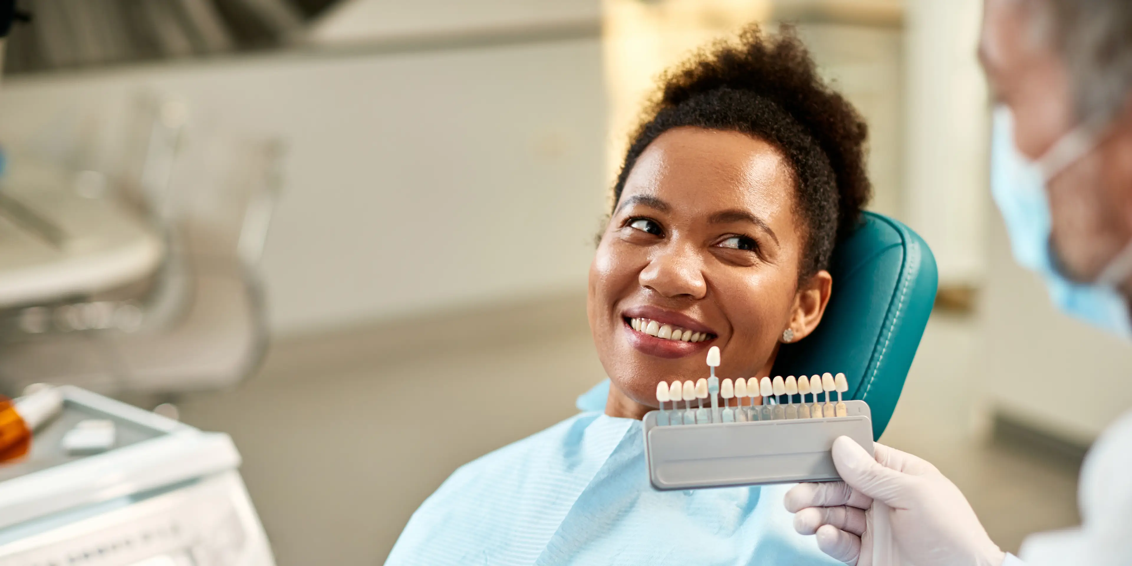 Smiling woman sitting in a dental chair holding a shade guide for teeth color matching with a dentist.