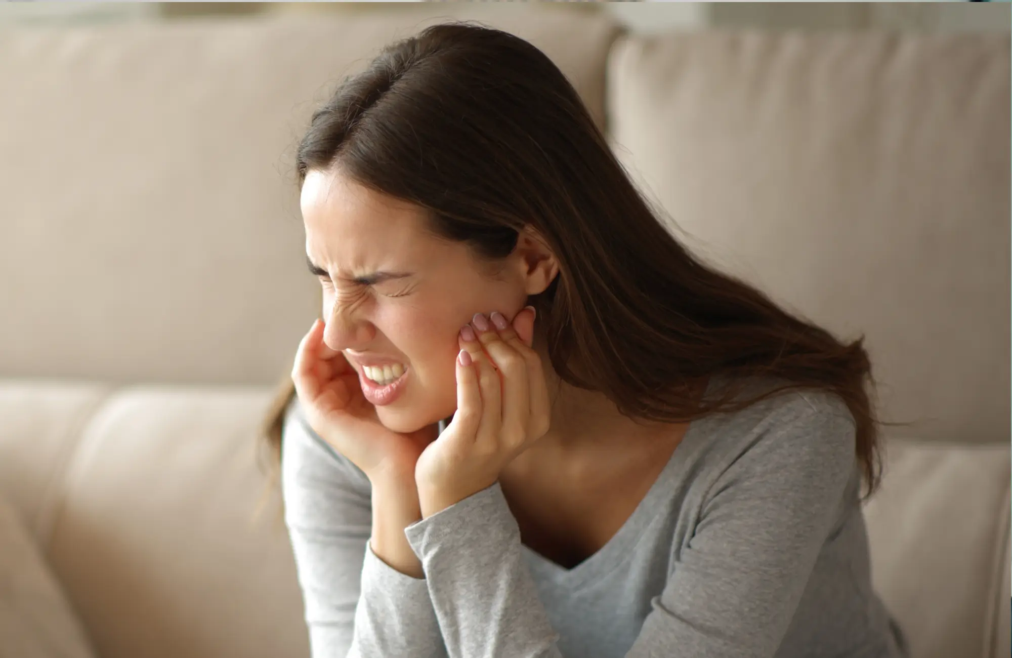 Woman sitting on a couch holding her jaw in pain with her eyes closed.