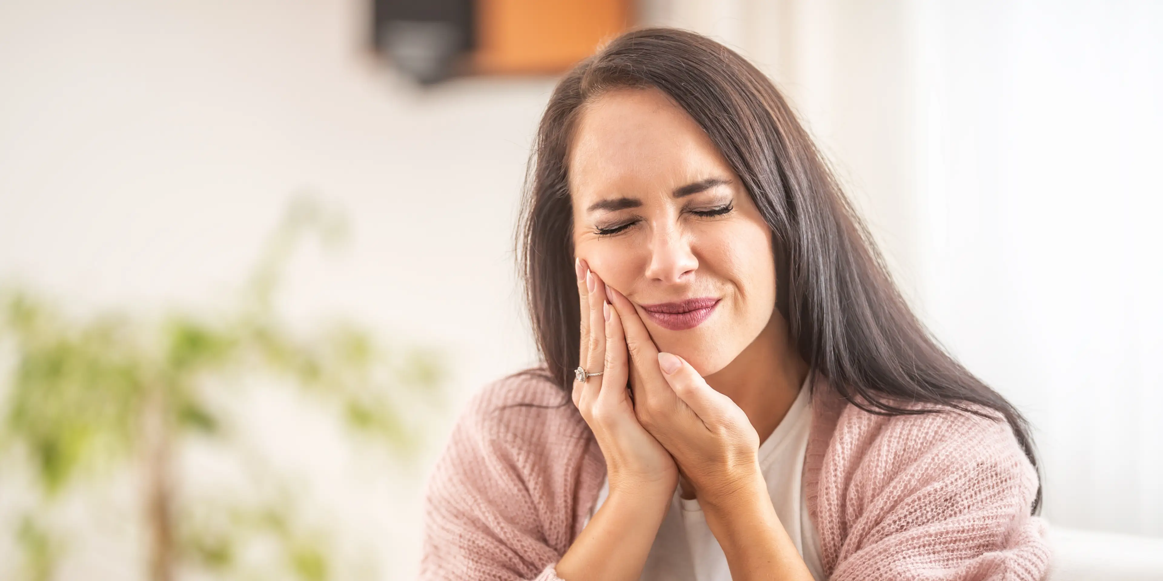 Woman with long dark hair holding her cheek in pain, wearing a pink sweater.
