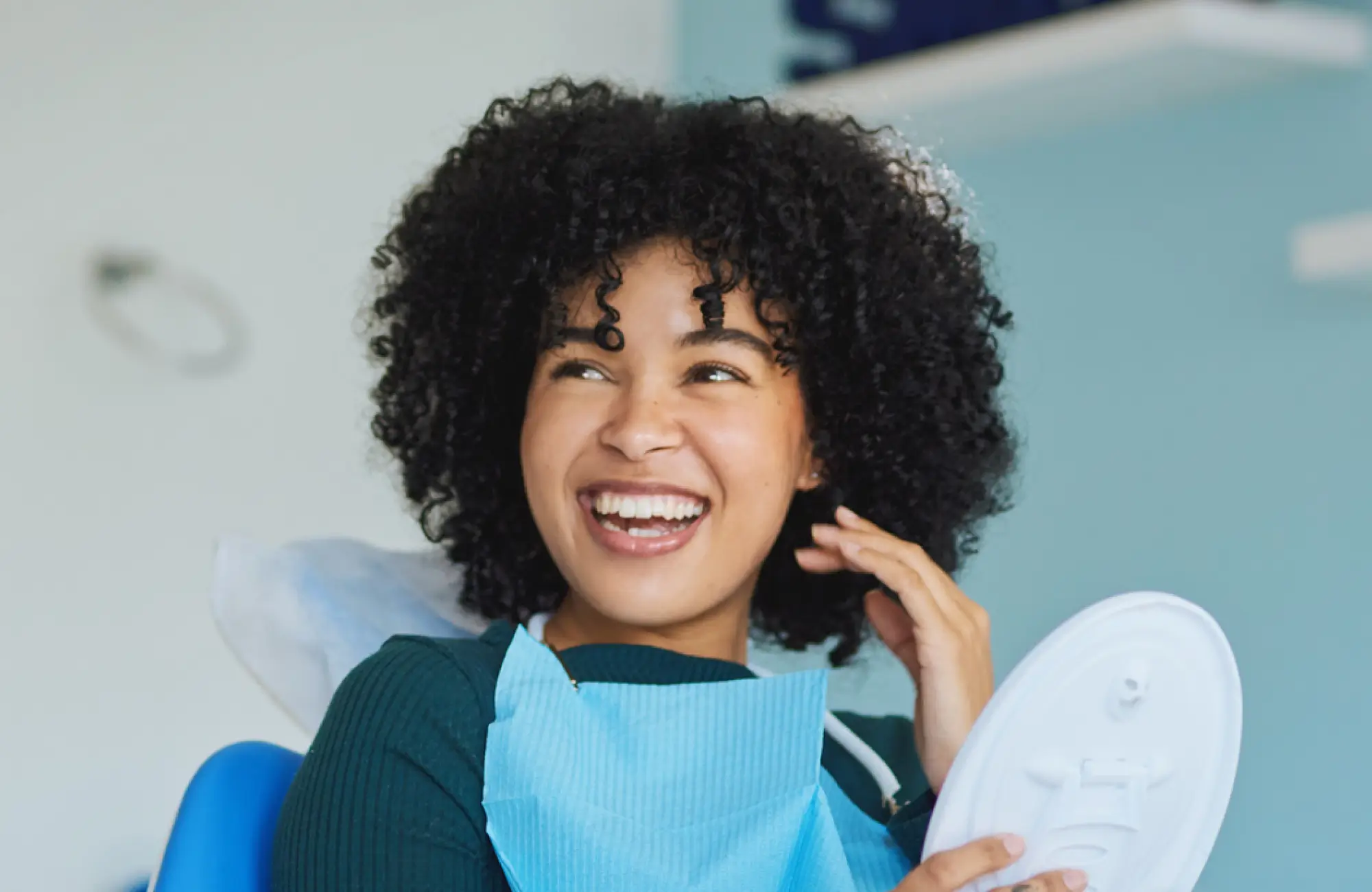 Smiling woman with curly hair in dental chair wearing a blue dental bib and holding a dental mirror.